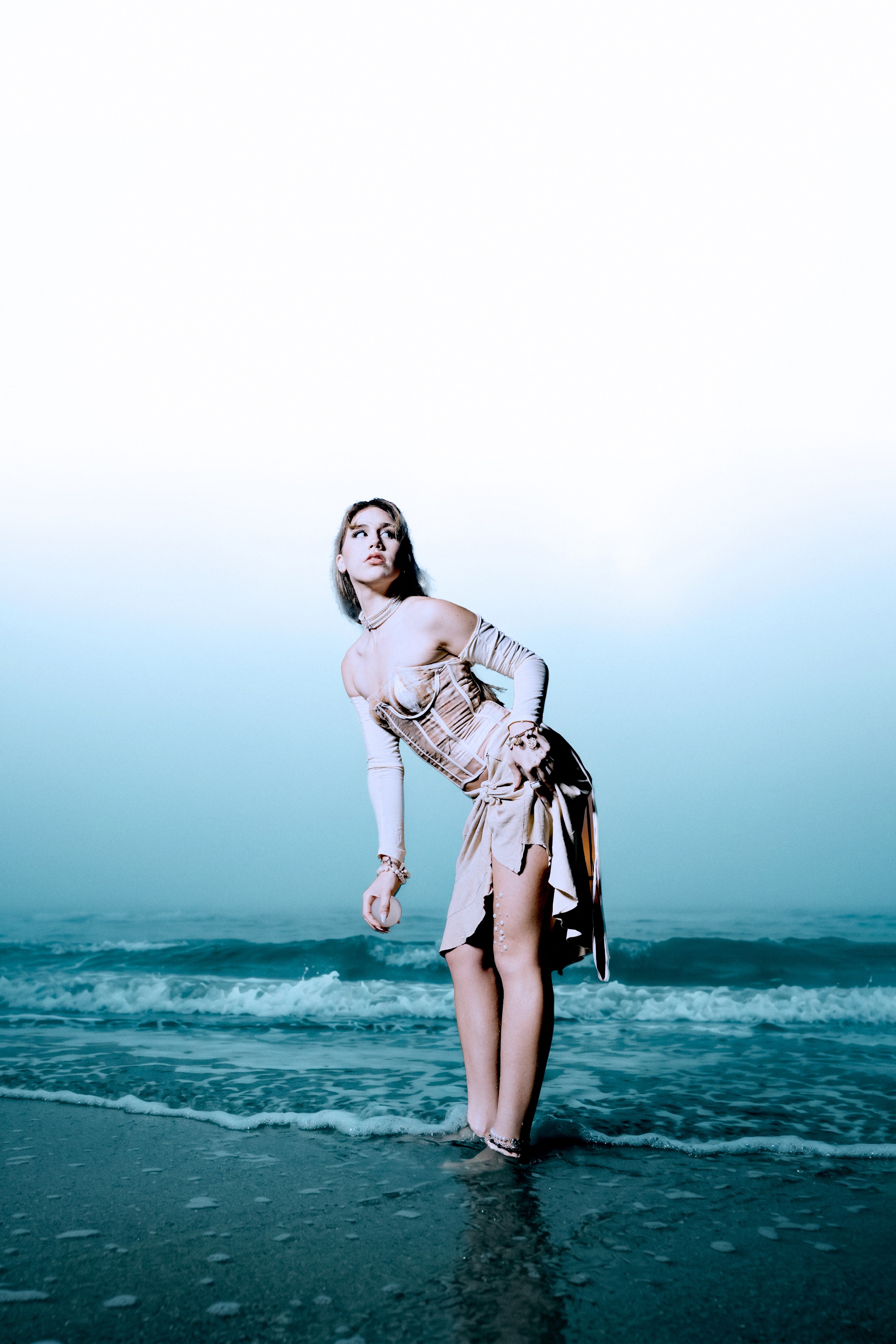 A fashion model stands at the shoreline in an ivory corset-inspired ensemble, looking upward as ocean waves roll behind her. A vast pale sky and cool blue tones frame the composition.
