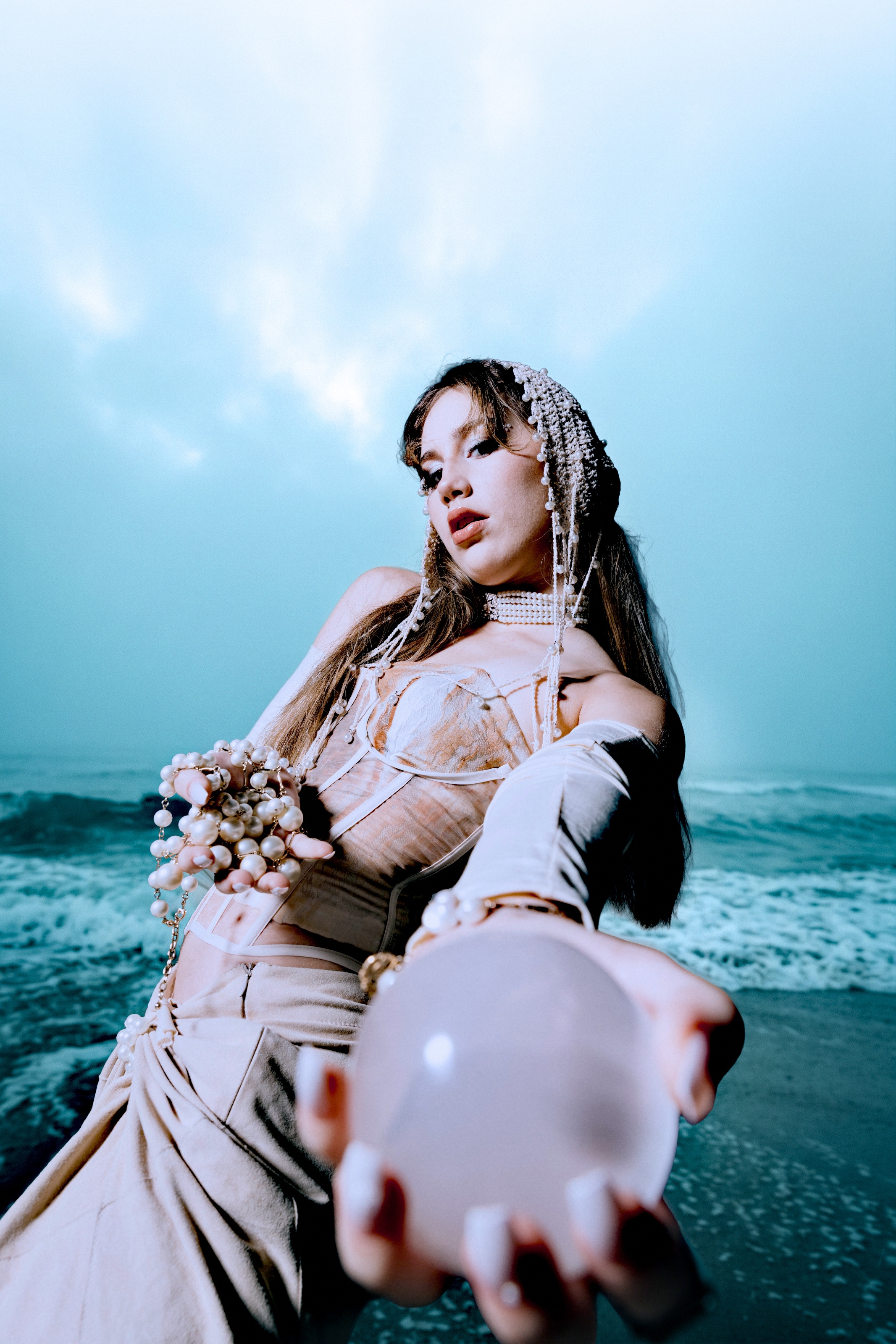 A low-angle fashion portrait of a model adorned with pearls, extending a translucent sphere toward the camera. Sculptural garments and layered jewelry contrast with the turquoise sea.
