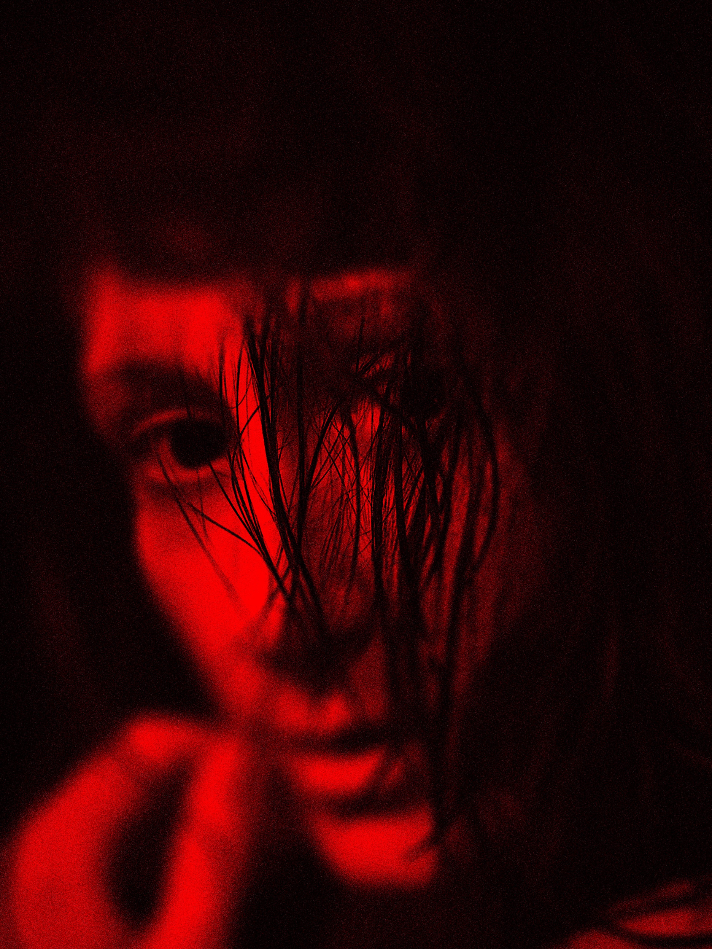 A portrait of a woman illuminated by intense red light. One eye emerges between dark strands of hair veiling part of her face and deep shadows. A black background reinforces the dramatic composition.