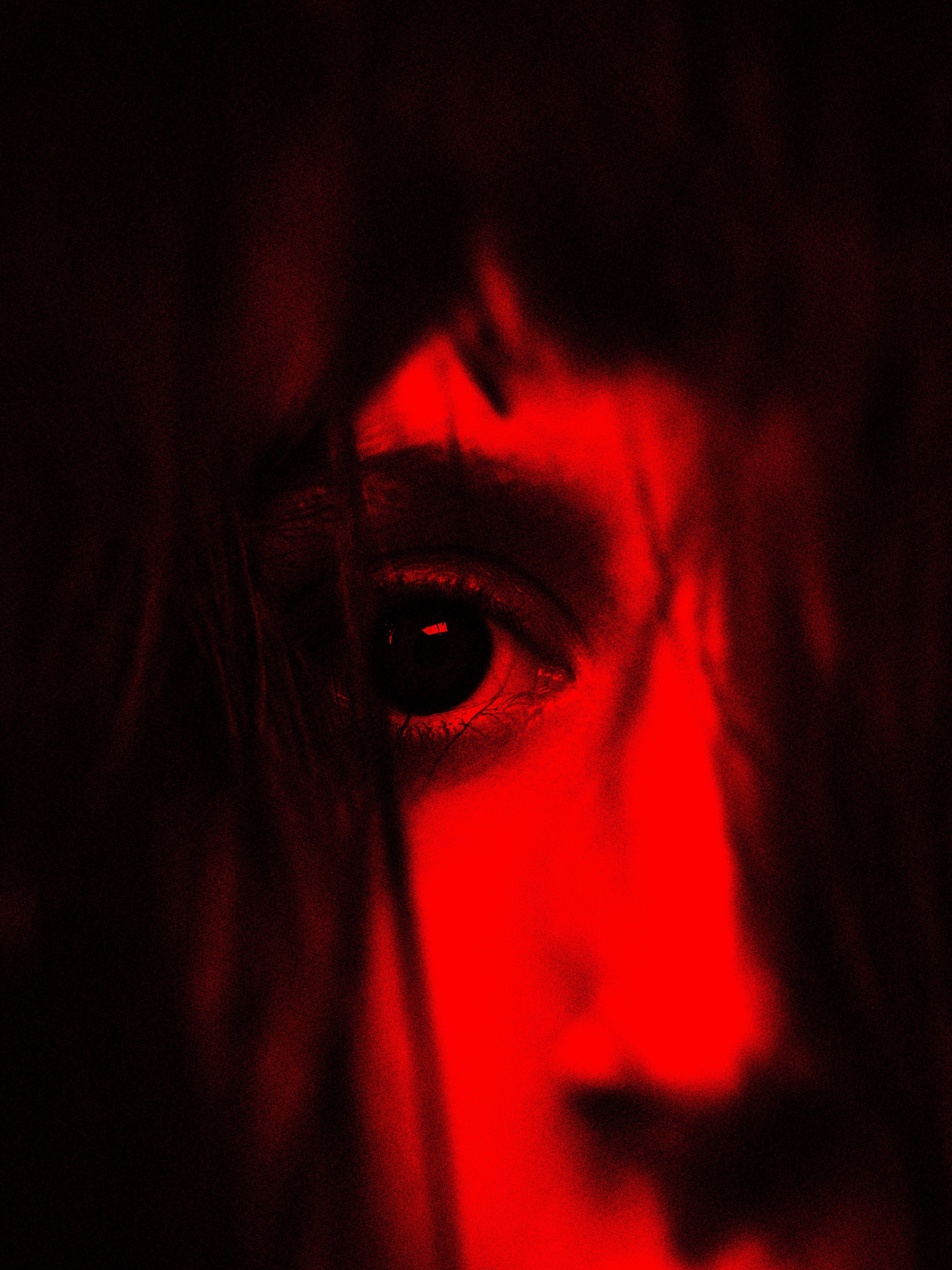A close-up portrait under red light, centered on a single eye. One side of the face remains in deep darkness, while red illumination on the other emphasizes the eye, nose and texture of the skin.